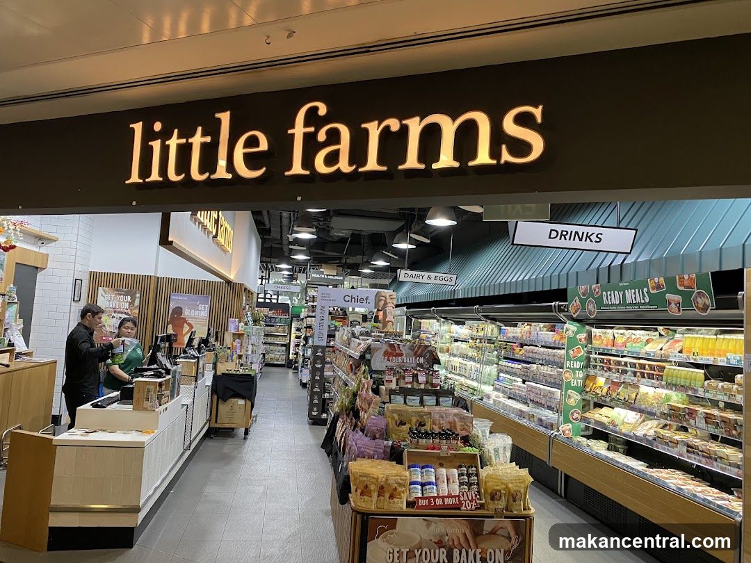 The exterior of a Little Farms store, with the prominent "little farms" logo above the entrance. Inside, shelves are stocked with various food items, including dairy, eggs, drinks, and ready meals. Two staff members are visible at a counter near the entrance.