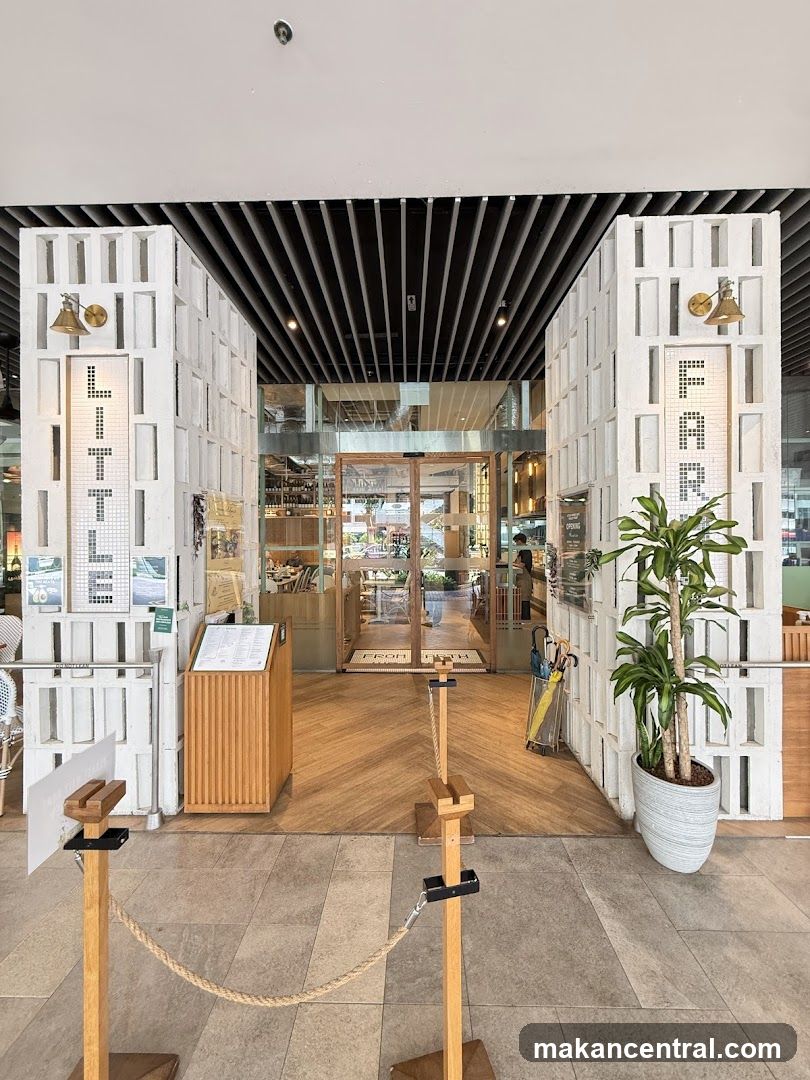 The entrance to a store called Little Farms. The store has a modern design with white brick pillars and a wooden floor. There are signs on the pillars that spell out "LITTLE FARMS". Inside the store, there are shelves stocked with products and tables for dining.