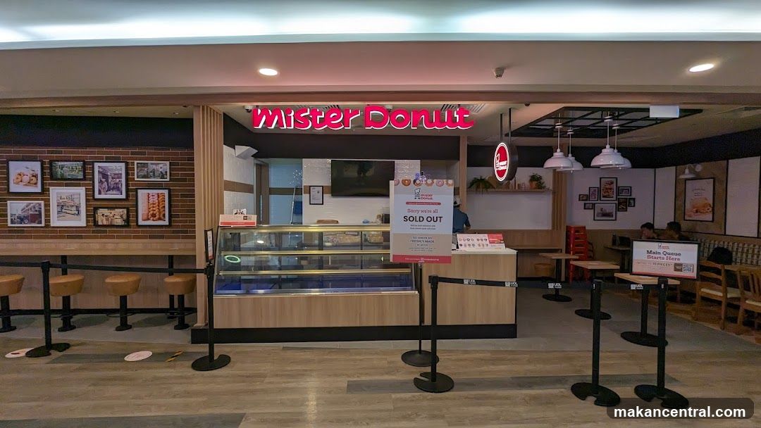 The exterior of a Mister Donut shop with a prominent neon sign. The shop features a display counter with empty shelves, a brick-patterned wall adorned with framed pictures, and seating areas. A "SOLD OUT" sign is displayed at the counter.