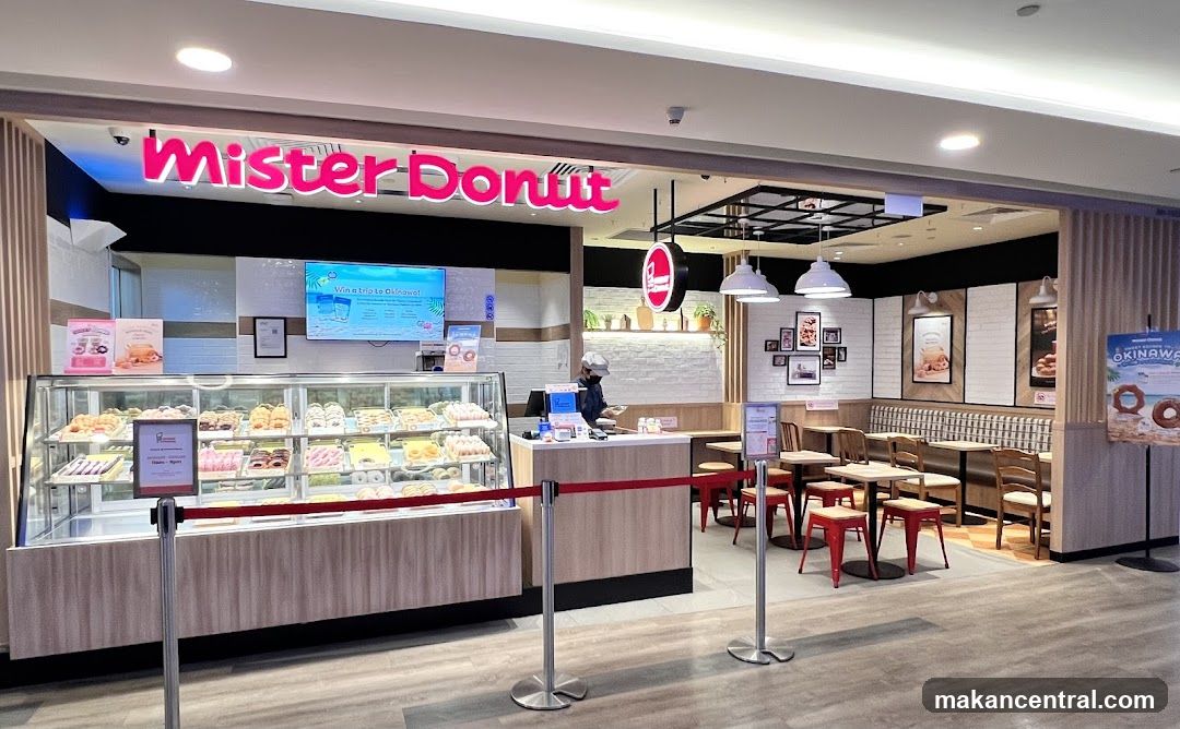 The exterior of a Mister Donut shop with a prominent pink neon sign above the entrance. A display case filled with various donuts is visible to the left, and seating areas with tables and chairs are arranged inside. A digital screen displays a promotion for a trip to Okinawa.