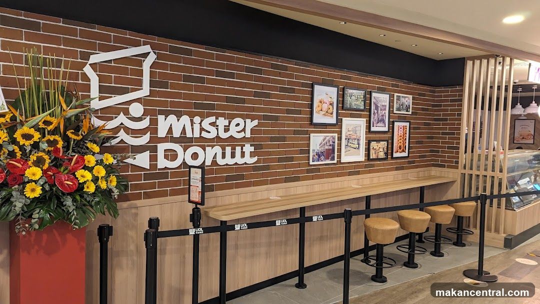 A Mister Donut storefront with a brick wall interior. The store's logo is prominently displayed in white lettering on the brick wall. A large floral arrangement with sunflowers and red anthuriums is placed to the left of the logo. Several framed pictures of donuts and street scenes are hung on the wall above a long wooden counter with stools. A display case with food items is visible to the right.