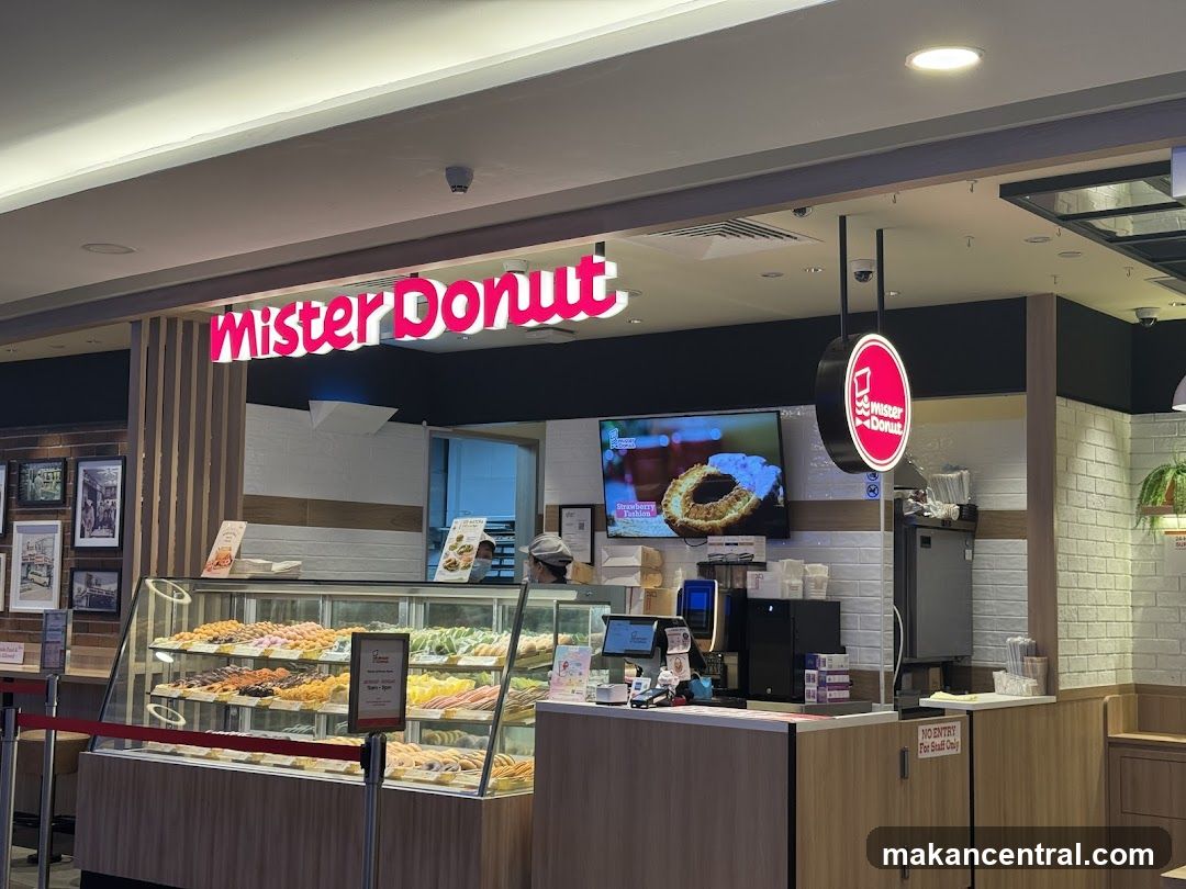 The storefront of a Mister Donut shop is visible, featuring a large illuminated sign that reads "Mister Donut" in pink lettering. Below the sign, a display case is filled with a variety of colorful donuts. A television screen mounted on the wall shows an advertisement for a donut. A circular hanging sign with the Mister Donut logo is also present. The interior of the shop, including a counter and staff members, is partially visible through the open entrance.
