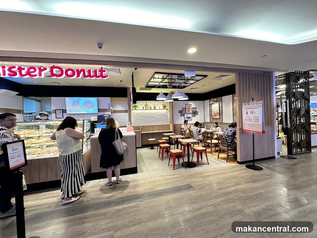 A view of the Mister Donut store entrance and interior. The prominent "Mister Donut" sign is visible above the counter. Inside, customers are seen ordering and dining at tables. The display case is filled with donuts. A sign indicating purchase limits is posted near the entrance.