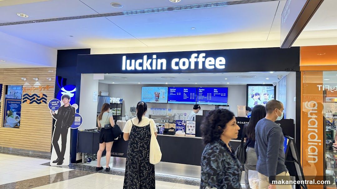 A Luckin Coffee store with its prominent signage above the counter. Customers are seen queuing and ordering. A cardboard cutout of a person is placed to the left of the entrance.