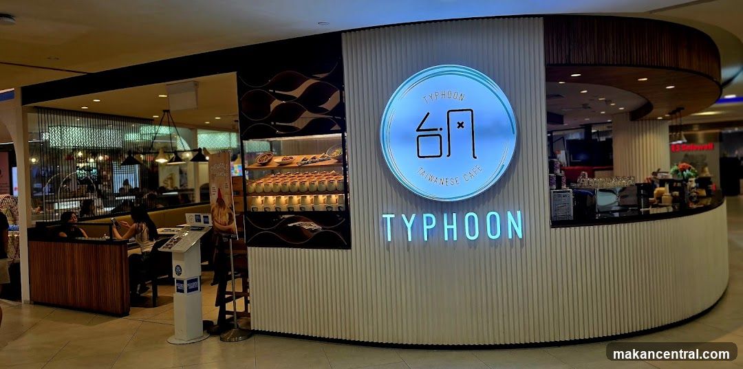 The exterior of a Taiwanese cafe named Typhoon. The cafe has a modern design with a curved counter and a prominent circular logo with the name "Typhoon" and Chinese characters. Below the logo, the name "TYPHOON" is displayed in large, illuminated letters. A display case shows rows of desserts. The interior of the cafe is visible, with seating areas and customers.