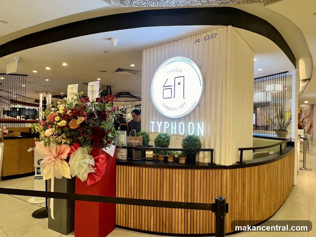 The exterior of a Typhoon Taiwanese Cafe with a prominent circular logo and neon sign. A large floral arrangement is placed to the left of the entrance.