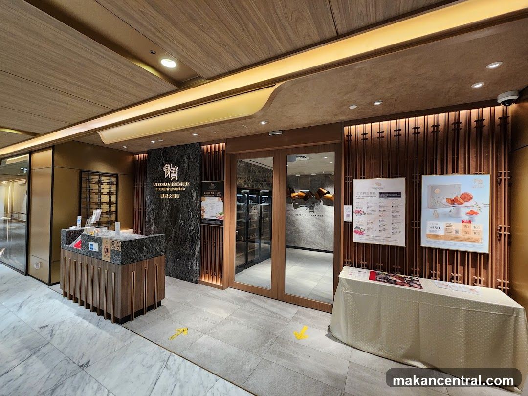 The entrance to Imperial Treasure Restaurant features a modern and elegant design with wooden accents and marble flooring. The restaurant's name is prominently displayed on a dark marble facade above the entrance. Glass doors lead into the interior, and to the right of the entrance, there are two posters displayed on the wall. One poster appears to be a menu, and the other advertises a product with pricing information. A table covered with a patterned cloth is placed in front of the posters.