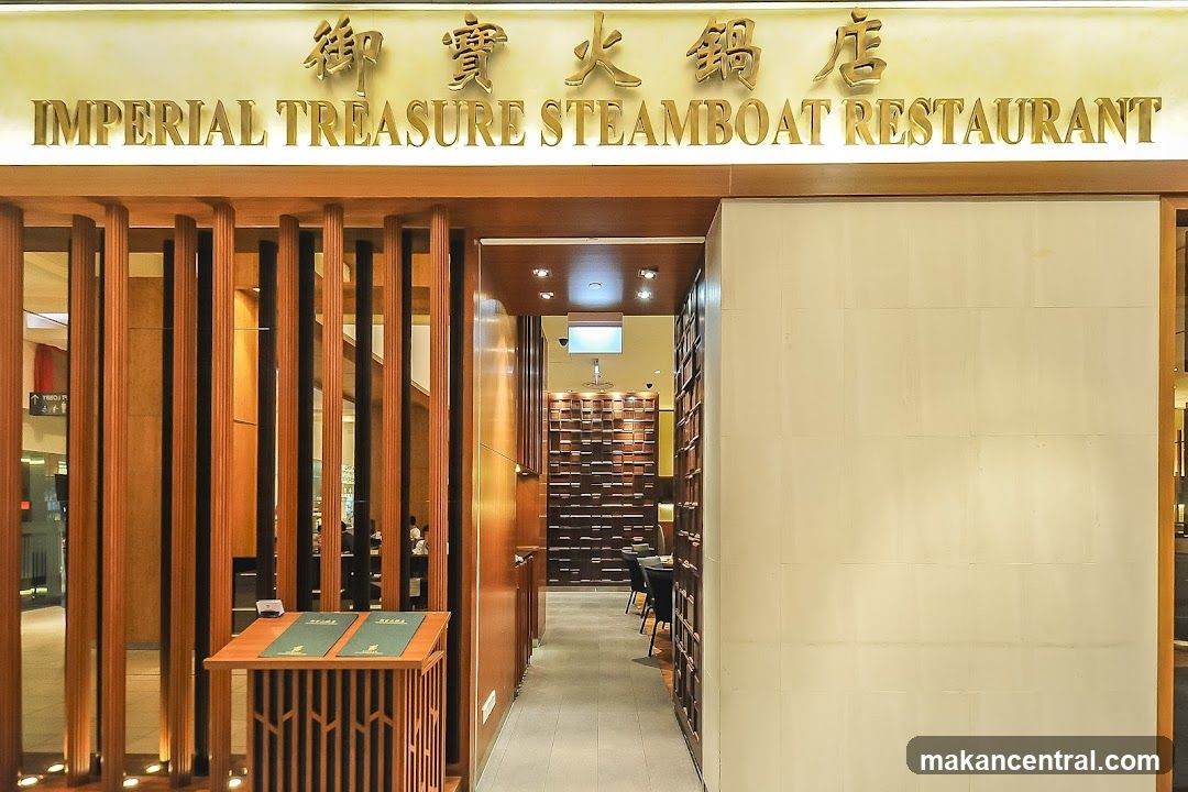 The exterior entrance of Imperial Treasure Steamboat Restaurant features a modern design with wooden accents. Above the entrance, the restaurant's name is displayed in prominent gold lettering, both in English and Chinese characters. The entryway is framed by vertical wooden slats, creating a warm and inviting atmosphere. Inside, a hallway leads to the dining area, which is visible in the background. A wooden stand with menus is placed near the entrance.