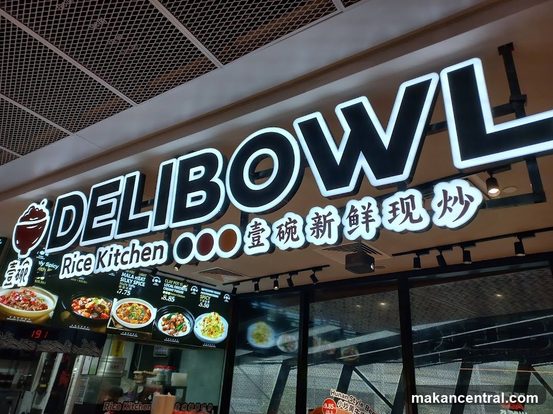 A brightly lit sign for DELIBOWL Rice Kitchen hangs above a food display. The sign features the restaurant name in large white letters with a black outline. Below the name, there are Chinese characters and the words "Rice Kitchen". A menu board is visible below the sign, displaying various dishes with images and prices.
