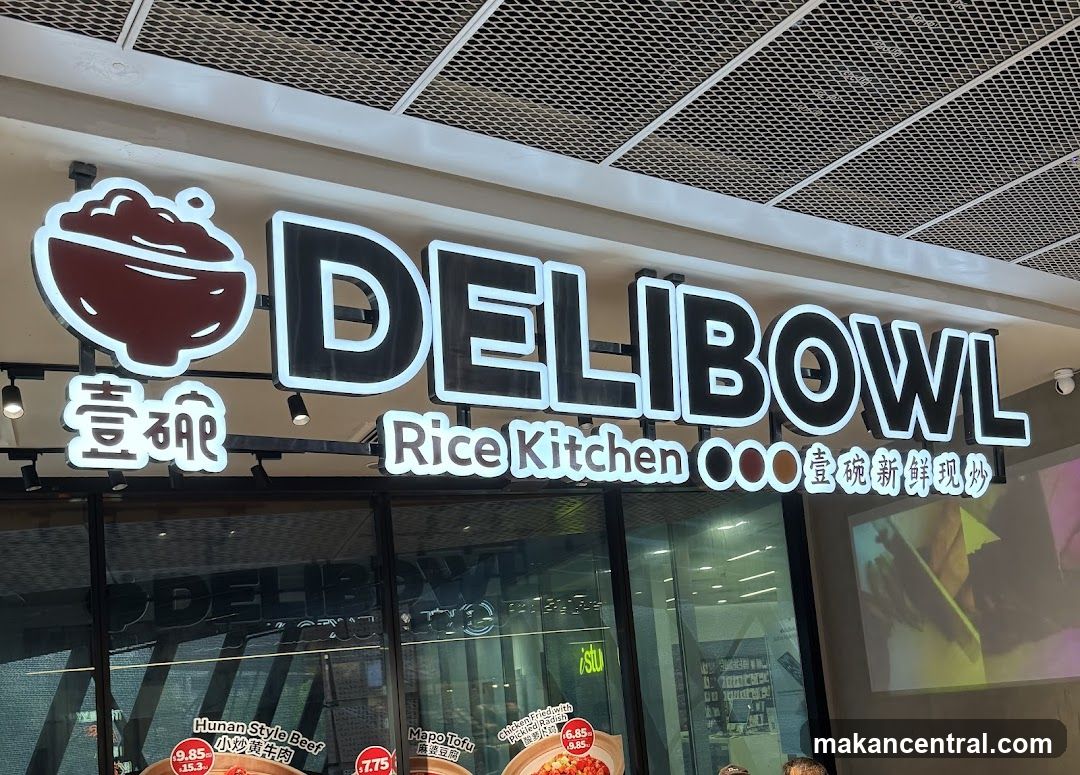A brightly lit sign for DELIBOWL Rice Kitchen is displayed above the storefront. The sign features the text "DELIBOWL" in large, bold letters with a white outline, accompanied by a graphic of a bowl of food. Below this, "Rice Kitchen" is written in smaller text, followed by Chinese characters. The storefront windows reflect the interior and the sign above. Visible menu items on display include "Hunan Style Beef" for 19.85 and "Mapo Tofu" for 6.85.