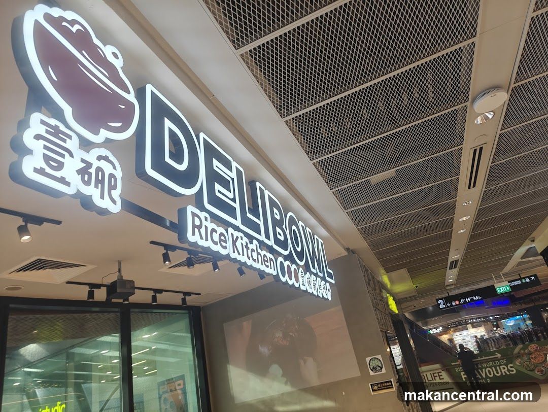 A brightly lit sign for Deli Bowl Rice Kitchen is visible on the exterior of a building. The sign features the words "DELI BOWL" in large white letters with a black outline, and below it, "Rice Kitchen" in smaller white letters. To the left of the main text is a graphic of a bowl with food and some Asian characters. The sign is illuminated and appears to be mounted above a storefront.