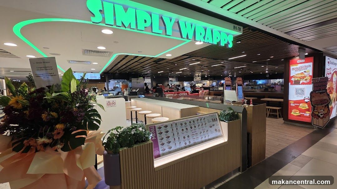 The exterior of a Simply Wrapps store with a prominent green neon sign. In the foreground, there is a display with a menu board and plants. To the right, there are promotional posters for burgers and drinks. The store appears to be located in a mall or food court.