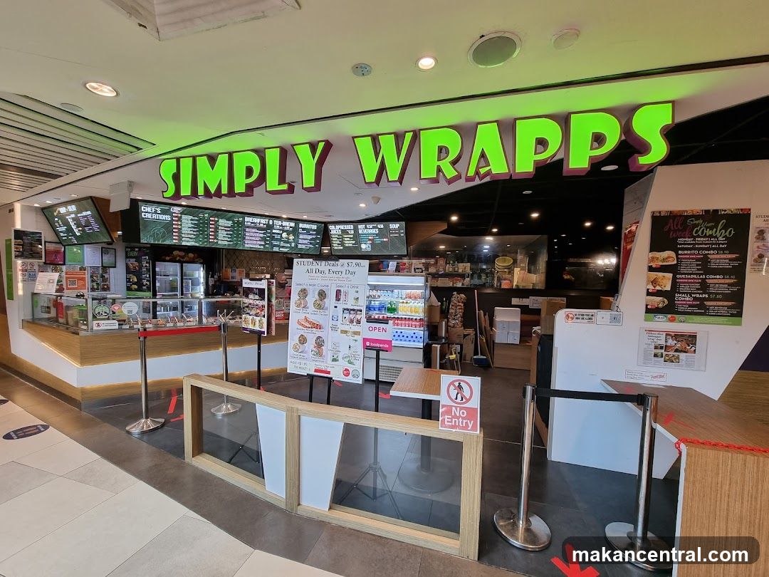 The exterior of a food establishment with a prominent green neon sign that reads "SIMPLY WRAPPS". The storefront features a counter with food displays, menu boards, and refrigerators. There are also promotional posters and signs visible.