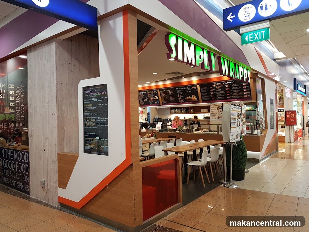 The exterior of a Simply Wrapps restaurant with its prominent green signage. The restaurant has a modern design with wooden accents and a glass facade. Inside, tables and chairs are set up for dining, and staff can be seen preparing food behind the counter. A menu board is visible above the counter, displaying various wrap options and chef's creations. A separate digital menu board is also present near the entrance.