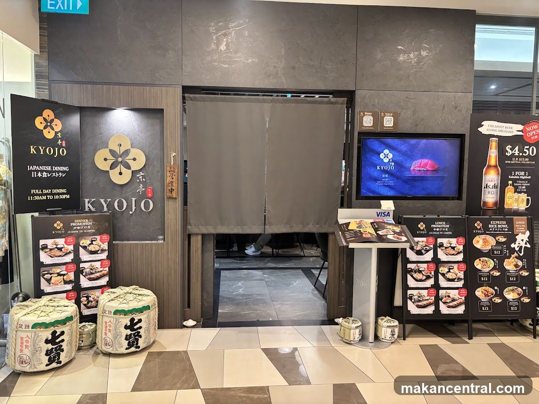 The exterior of a Japanese restaurant called Kyojo. A large sign on the left displays the restaurant name, "KYOJO JAPANESE DINING," and operating hours. A curtain hangs over the entrance. To the right, a television screen shows the Kyojo logo and an image of sushi. Promotional posters for lunch and beer specials are displayed. Two large decorative barrels are placed in front of the entrance.