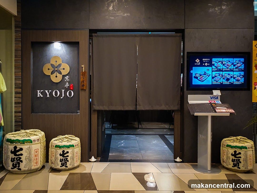 The entrance to a restaurant called KYOJO. A large golden emblem is displayed above the name. To the right of the entrance, a digital screen displays menu items. Several large sake barrels are placed in front of the entrance.