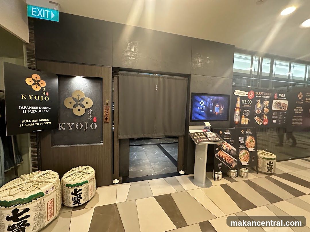 The exterior of a Japanese restaurant called Kyojo. A black sign to the left displays the restaurant name, "Kyojo," "Japanese Dining," and "Full Day Dining" with operating hours from 11:30 AM to 10:30 PM. The restaurant entrance is marked by a dark wooden door with a curtain. To the right of the entrance, a digital screen displays the Kyojo logo and images of drinks. A stand in front of the screen shows a menu with various sushi and food items. Two large decorative barrels are placed in front of the establishment.