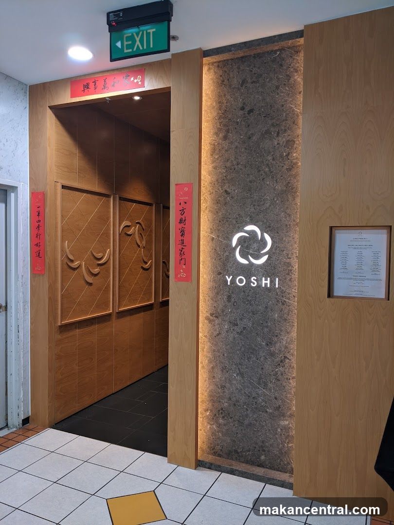 The entrance to a restaurant with a wooden facade and a central panel featuring the name "YOSHI" and a circular logo. Red Chinese calligraphy banners are affixed to the wooden pillars. An "EXIT" sign is visible above the entrance. A framed document is mounted on the right side of the facade.