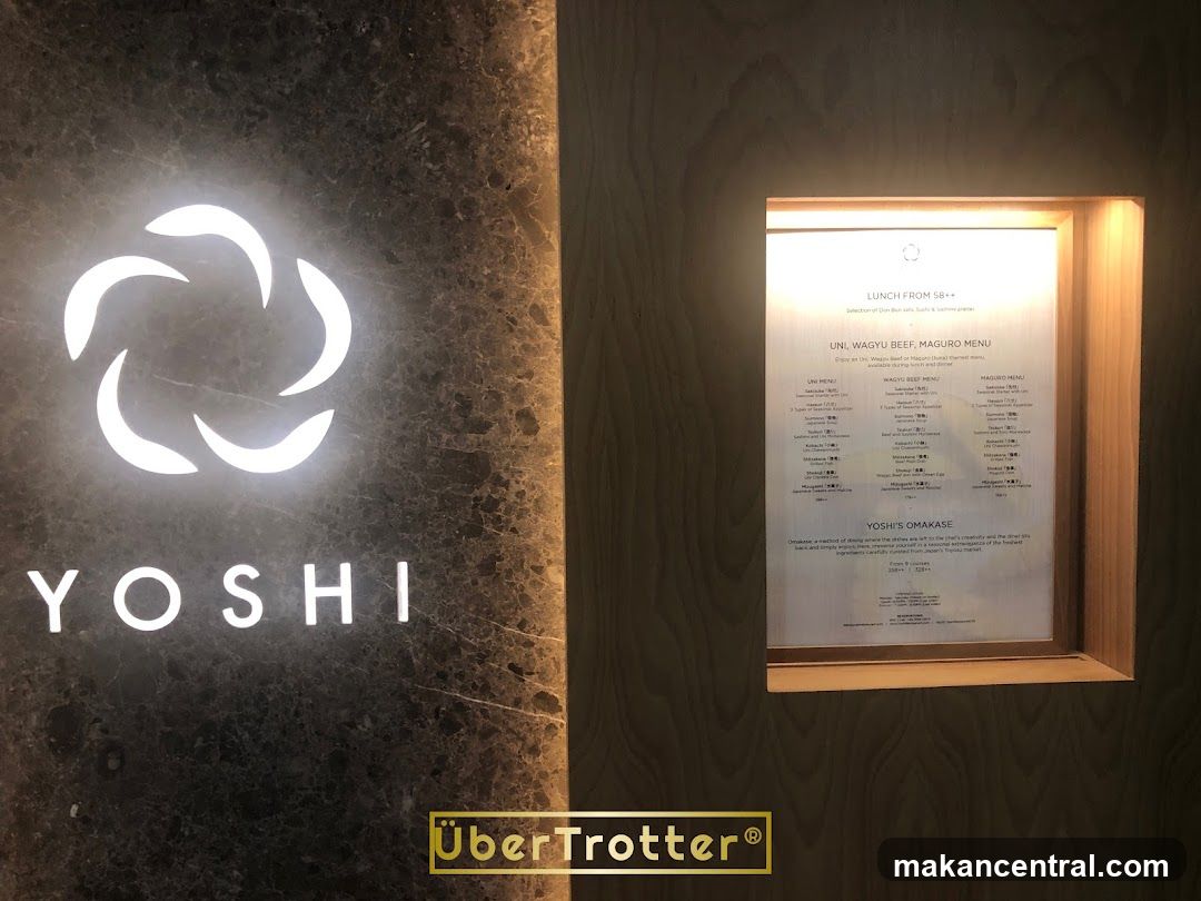 A close-up of a restaurant's exterior signage and a menu displayed in a wooden frame. The signage features a white, abstract circular logo above the illuminated white text "YOSHI". To the right, a menu is visible within a recessed wooden panel, detailing lunch options including Uni, Wagyu Beef, and Maguro menus, as well as Yoshi's Omakase.