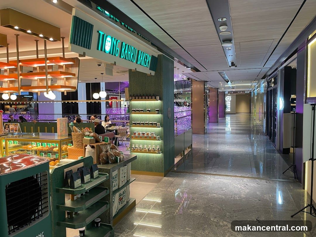 The exterior of Tiong Bahru Bakery, a brightly lit establishment with a green and white color scheme. The bakery's name is prominently displayed in neon green lettering above the entrance. Inside, display cases showcase an assortment of pastries and baked goods. Shelves are stocked with jars and packaged items. The interior features modern lighting and seating areas where customers are visible. The bakery is situated within a larger, modern shopping mall with polished floors and ambient lighting.