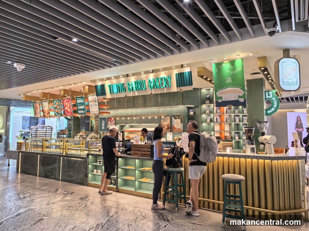 A view of the Tiong Bahru Bakery storefront with customers ordering at the counter. The bakery features a green and gold color scheme with display cases filled with pastries.