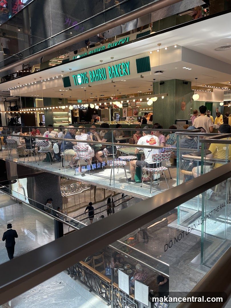 The exterior of Tiong Bahru Bakery, a brightly lit establishment with green accents and a prominent sign. People are seated at tables inside and on a mezzanine level, enjoying food and drinks. The bakery appears to be located within a modern shopping mall, with multiple levels visible.