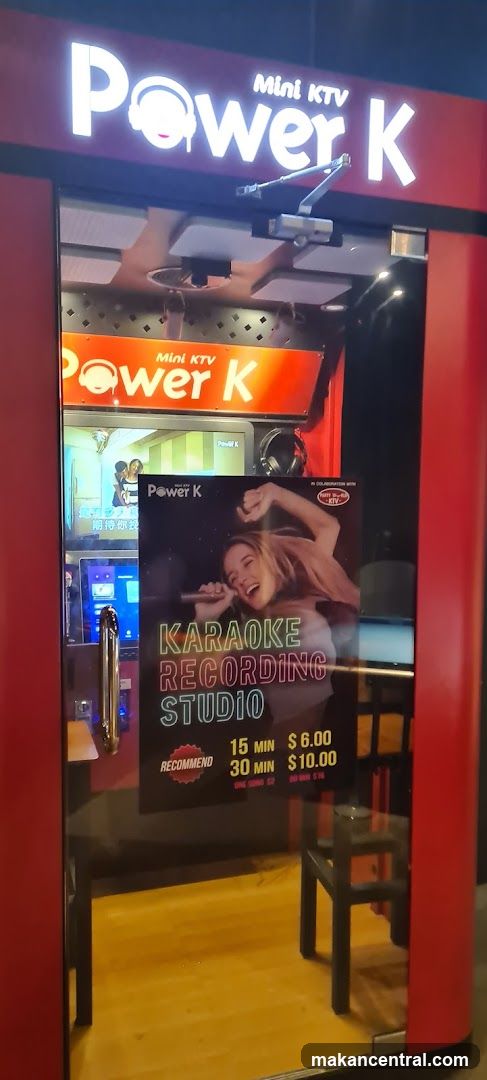 The exterior of a red and black karaoke booth with a glass door. The sign above the door reads "Power K Mini KTV" in white illuminated letters. A poster on the glass door advertises "Karaoke Recording Studio" with pricing for 15 minutes at $6.00 and 30 minutes at $10.00.