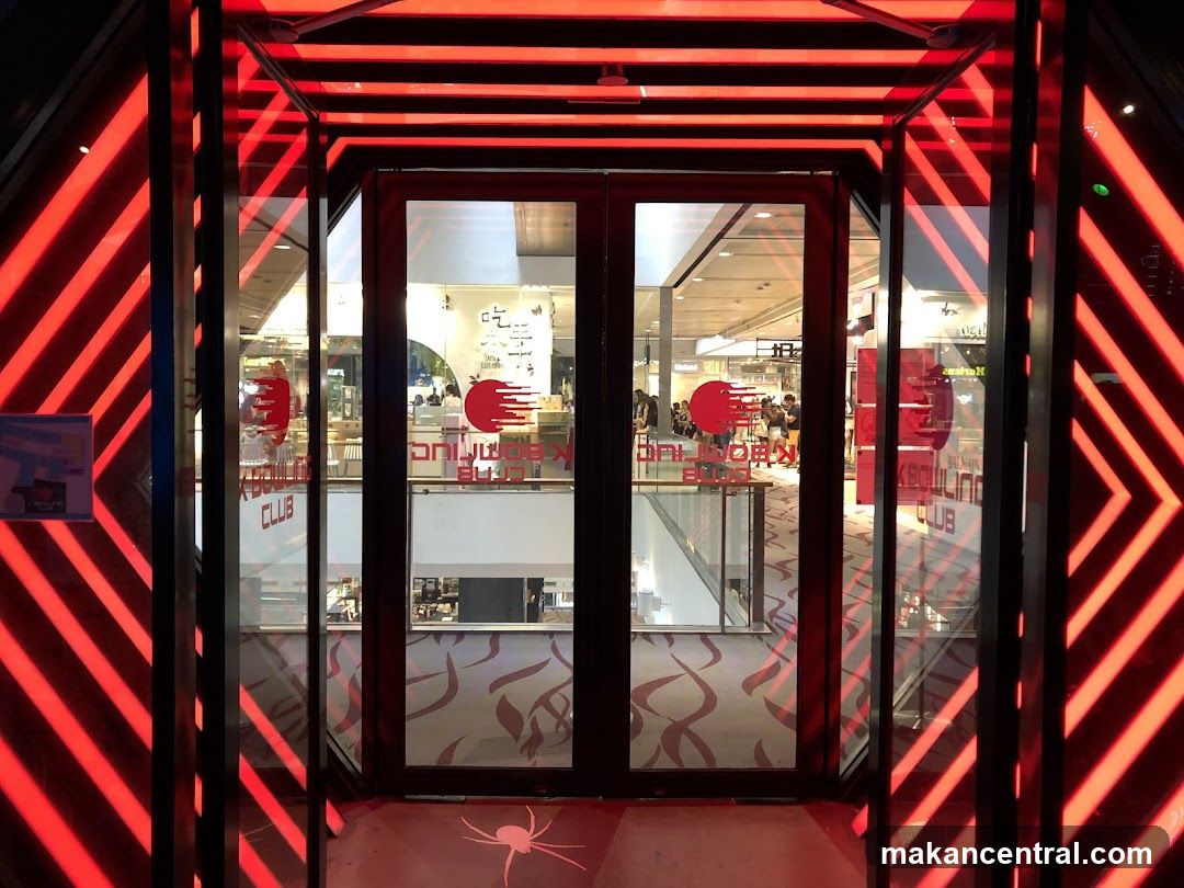 A modern club entrance with double glass doors framed by striking red neon lights. The lights create a geometric pattern on the walls and ceiling, leading into the interior. Through the glass doors, a brightly lit interior with people and tables is visible. The club's logo, a red circle with a horizontal line, is displayed on the glass doors along with text that appears to read "K-BOWLING CLUB". A spider graphic is projected onto the floor in front of the entrance.