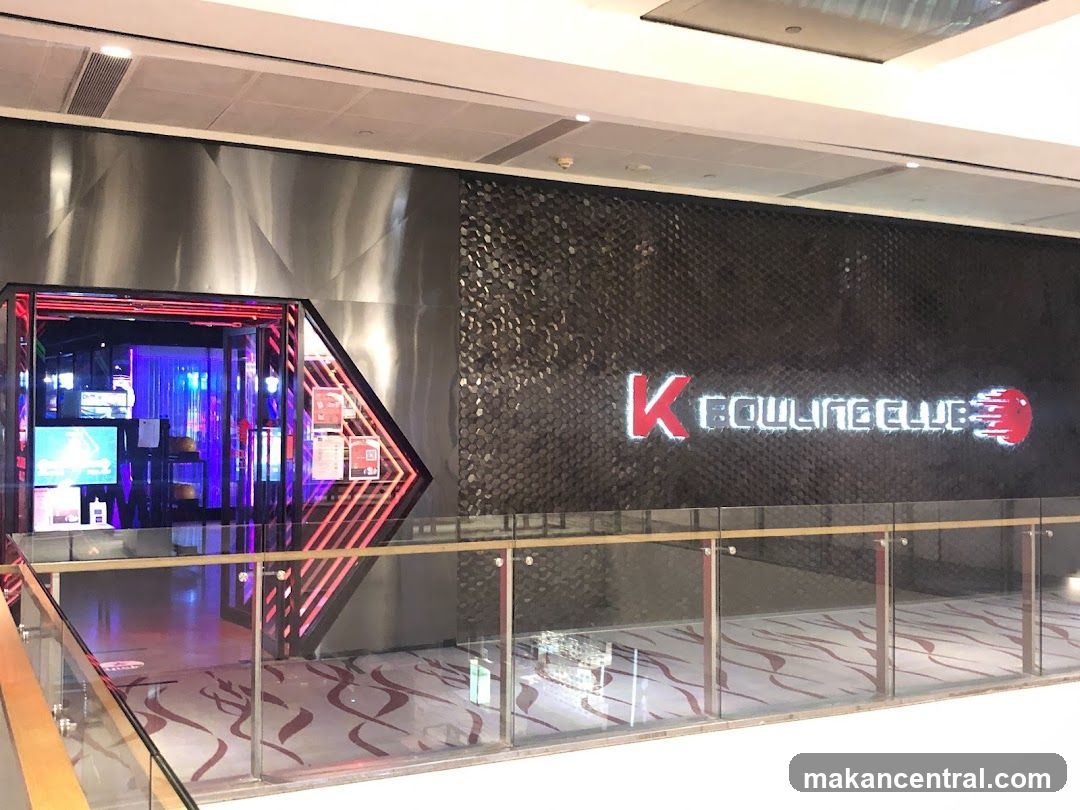 The entrance to K Bowling Club is visible, with a neon sign displaying the club's name and logo. The entrance is framed by red neon lights and has a modern, sleek design. Inside, colorful lights and screens suggest an entertainment venue.