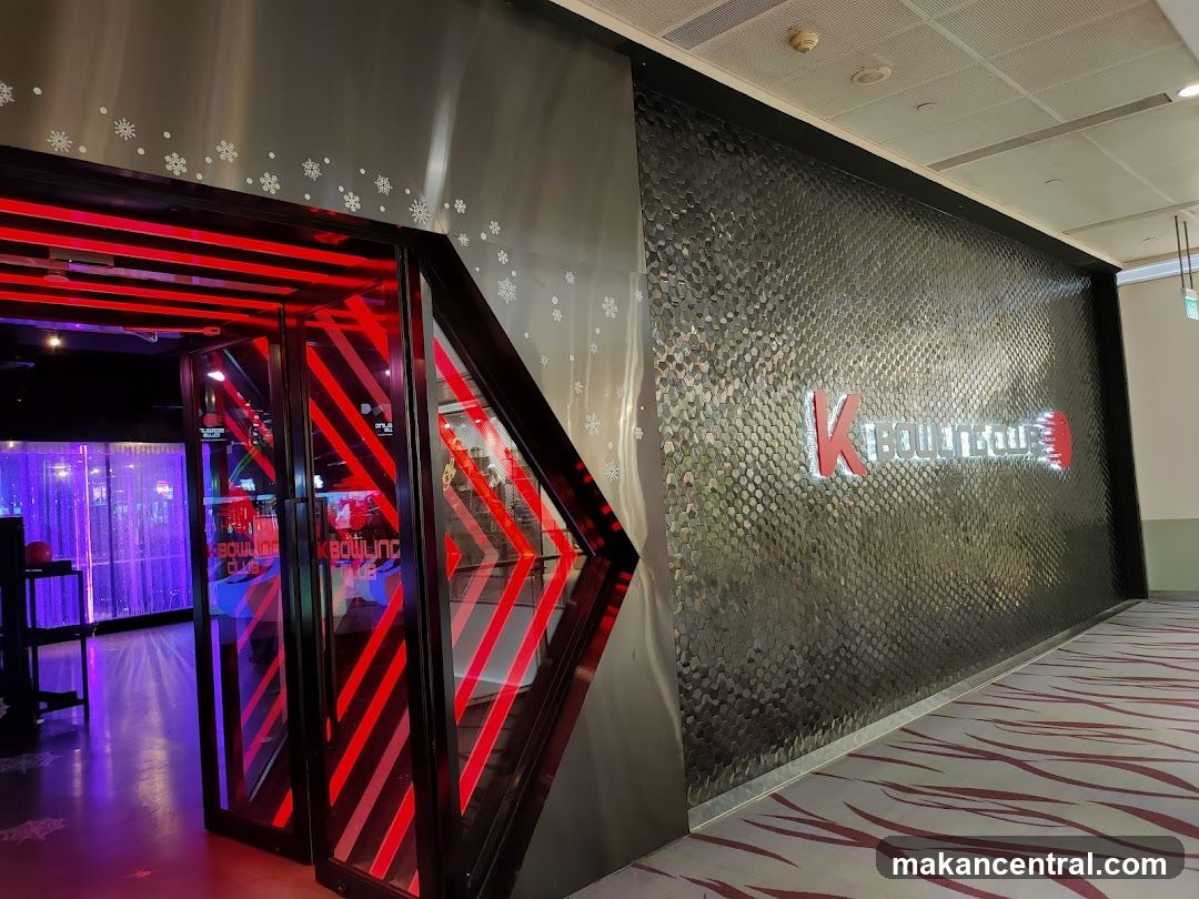 The entrance to K Bowling Club features a modern facade with a textured wall and illuminated signage. The doors are glass with red diagonal stripes, and the interior is visible, showing purple lighting and bowling lanes. Snowflakes are visible as a decorative element on the wall near the entrance.