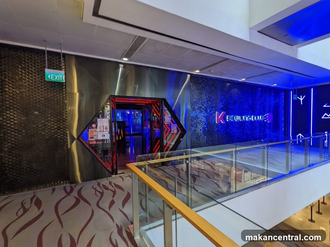 The entrance to K Bowling Club is visible, with a neon sign displaying the club's name. The entrance is framed by red neon lights and has a modern, sleek design. The surrounding wall features a blue, textured pattern and more neon light accents. An exit sign is also visible to the left.