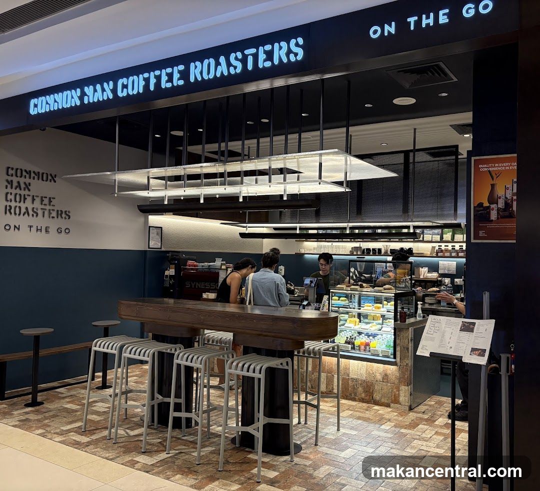 The exterior of a coffee shop with neon signage that reads "COMMON MAN COFFEE ROASTERS ON THE GO". There is a counter with stools and a display case with pastries.