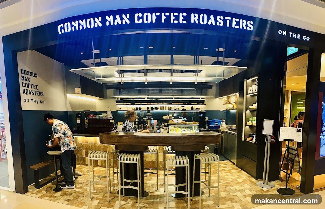 A coffee shop with a prominent sign reading "COMMON MAN COFFEE ROASTERS ON THE GO". The interior features a counter with bar stools, coffee machines, and display cases with food items. Two customers are visible, one ordering and another behind the counter.