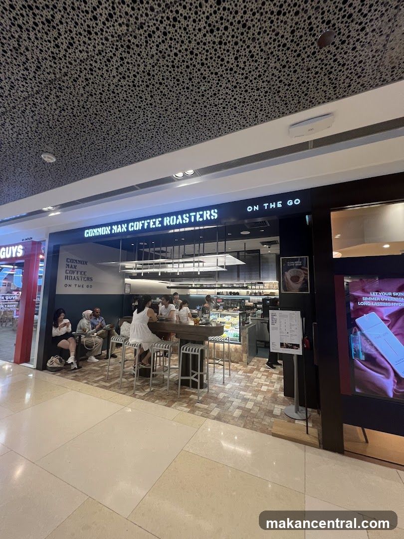 The exterior of a coffee shop with the name "COMMON MAN COFFEE ROASTERS ON THE GO" displayed prominently on the facade. Several people are seated at a counter inside the shop.