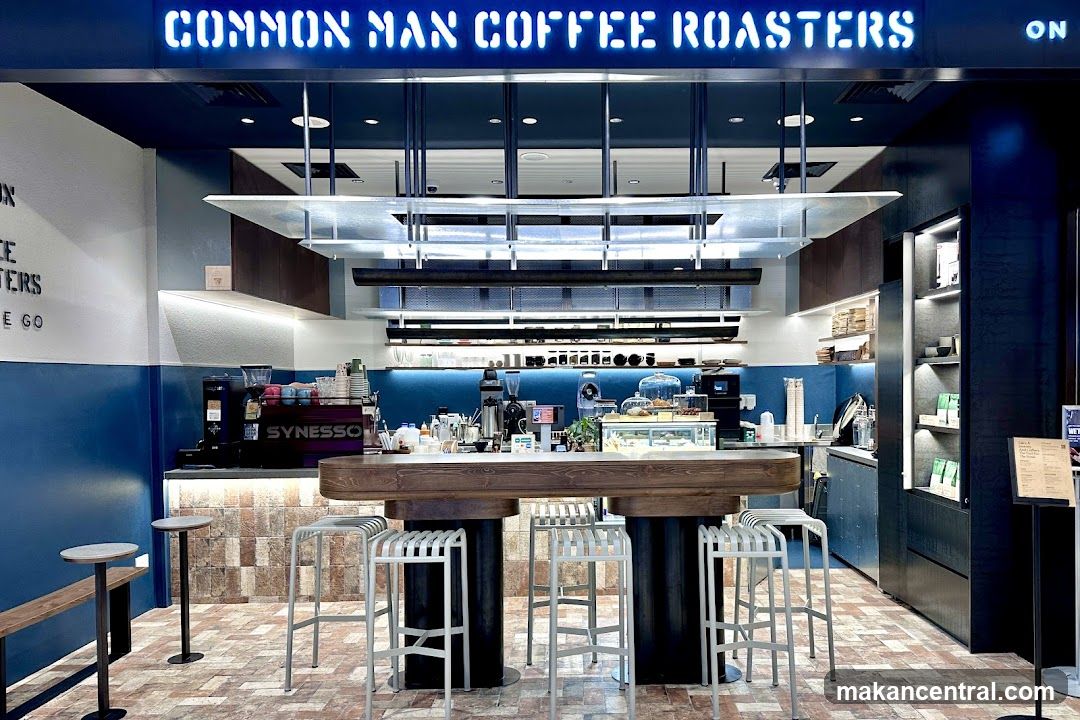 The exterior of a coffee shop with a neon sign that reads "COMMON MAN COFFEE ROASTERS". The interior features a long wooden counter with stools, a coffee machine, and display cases with pastries. The walls are painted blue and white, and the floor is tiled.