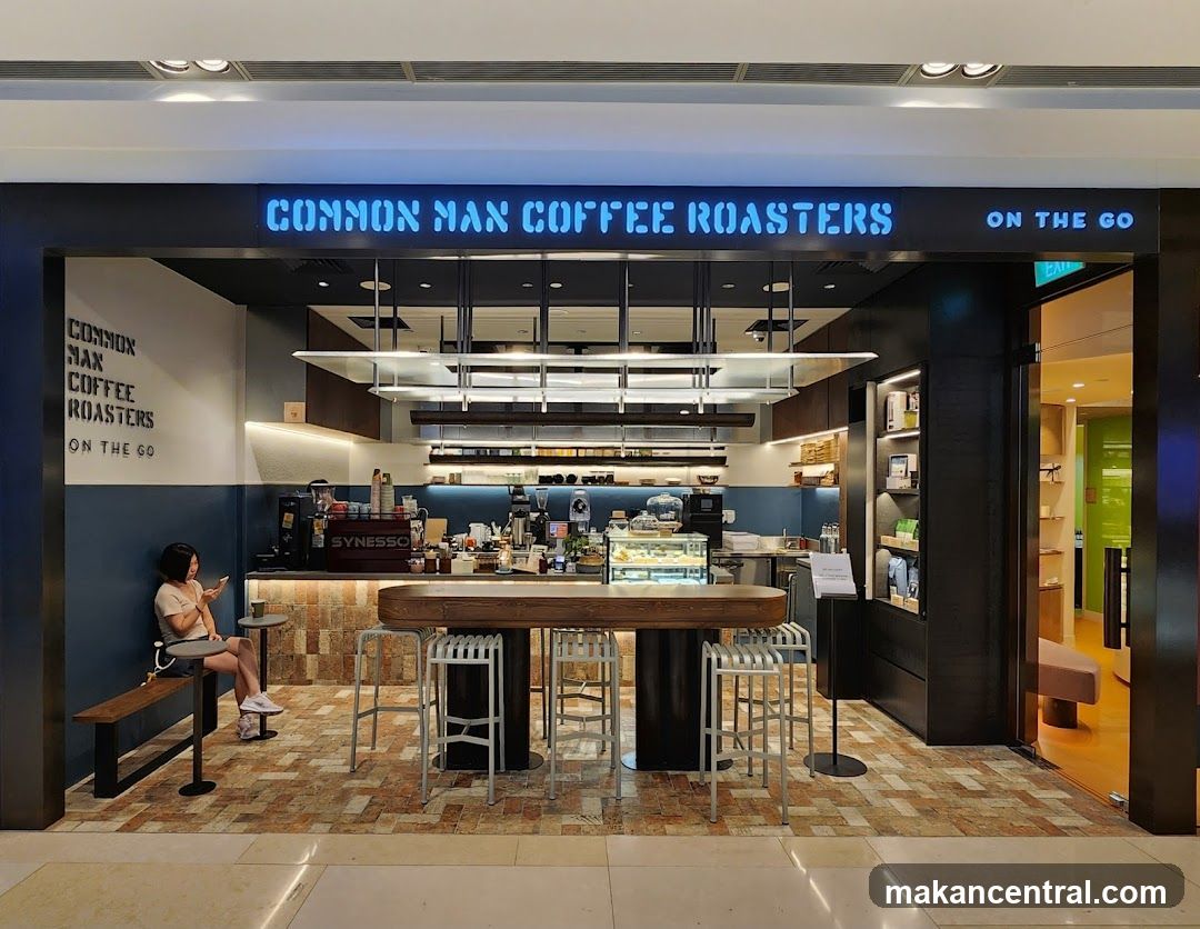 A brightly lit coffee shop with a prominent neon sign reading "COMMON MAN COFFEE ROASTERS ON THE GO". The interior features a long wooden counter with stools, a display case with pastries, and a coffee machine. A person is seated on a bench to the left, looking at their phone. The shop has a modern and clean aesthetic.