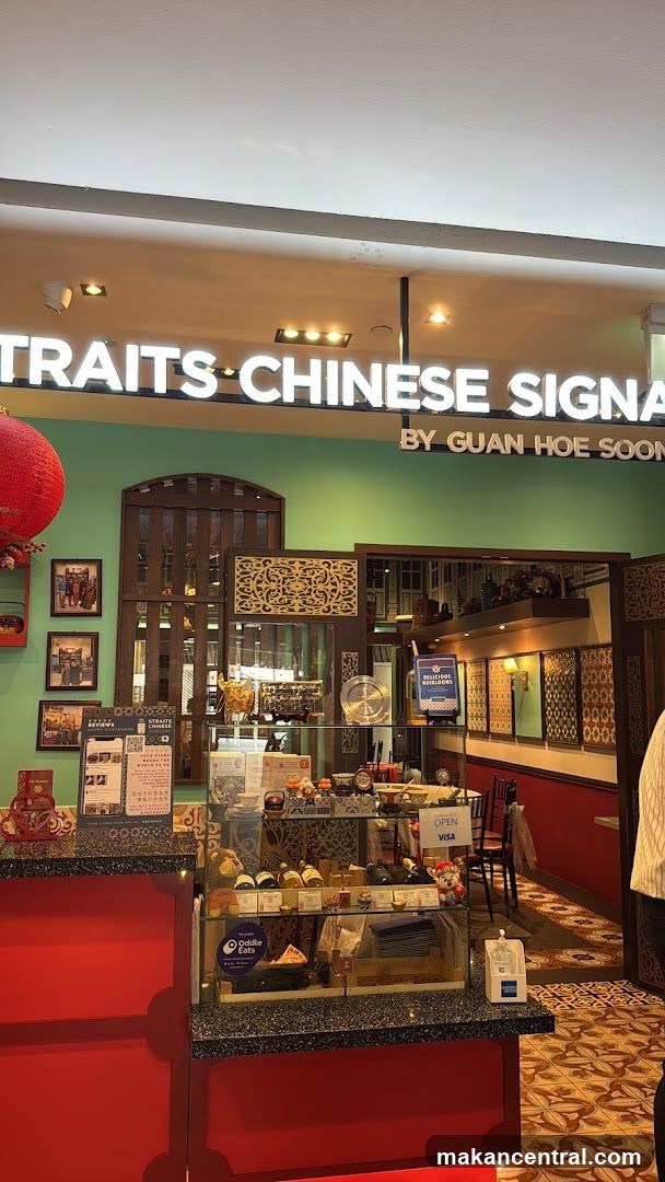 The exterior of a shop with a bright green wall and a large illuminated sign that reads "PORTRAITS CHINESE SIGNATURES BY GUAN HOE SOON". Below the sign, there is a display counter with various items, including bottles, jars, and what appear to be pastries or desserts. The shop has traditional wooden decorative elements and framed pictures on the wall. A red lantern hangs to the left. The floor has a patterned tile design.