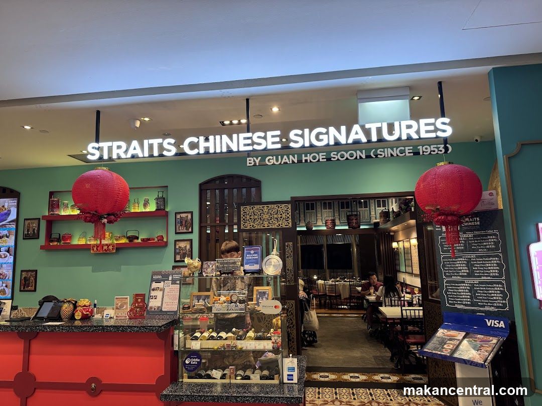The exterior of a restaurant with a green wall and a red counter. A large sign above reads "STRAITS CHINESE SIGNATURES BY GUAN HOE SOON (SINCE 1953)". Red lanterns hang near the entrance. Inside, tables and chairs are visible. A menu stand with a VISA logo is placed to the right of the entrance.