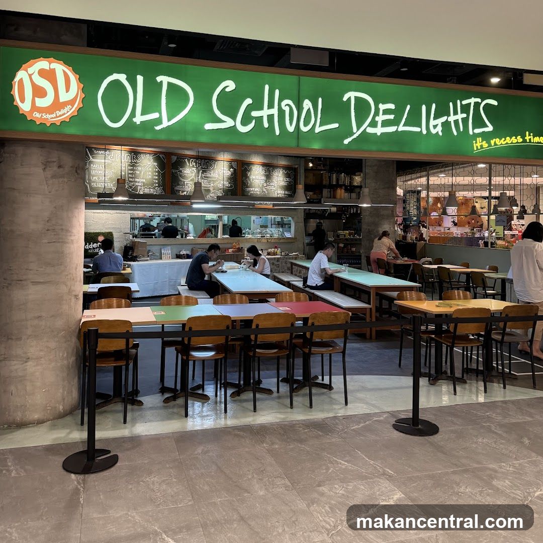 The exterior of a restaurant called Old School Delights. The restaurant has a large green sign with white lettering that says "OLD SCHOOL DELIGHTS" and a smaller logo on the left. Below the sign, there are menus displayed on chalkboards. Inside, there are several tables and chairs, and people are seated and eating. The restaurant appears to be in a food court or mall setting.
