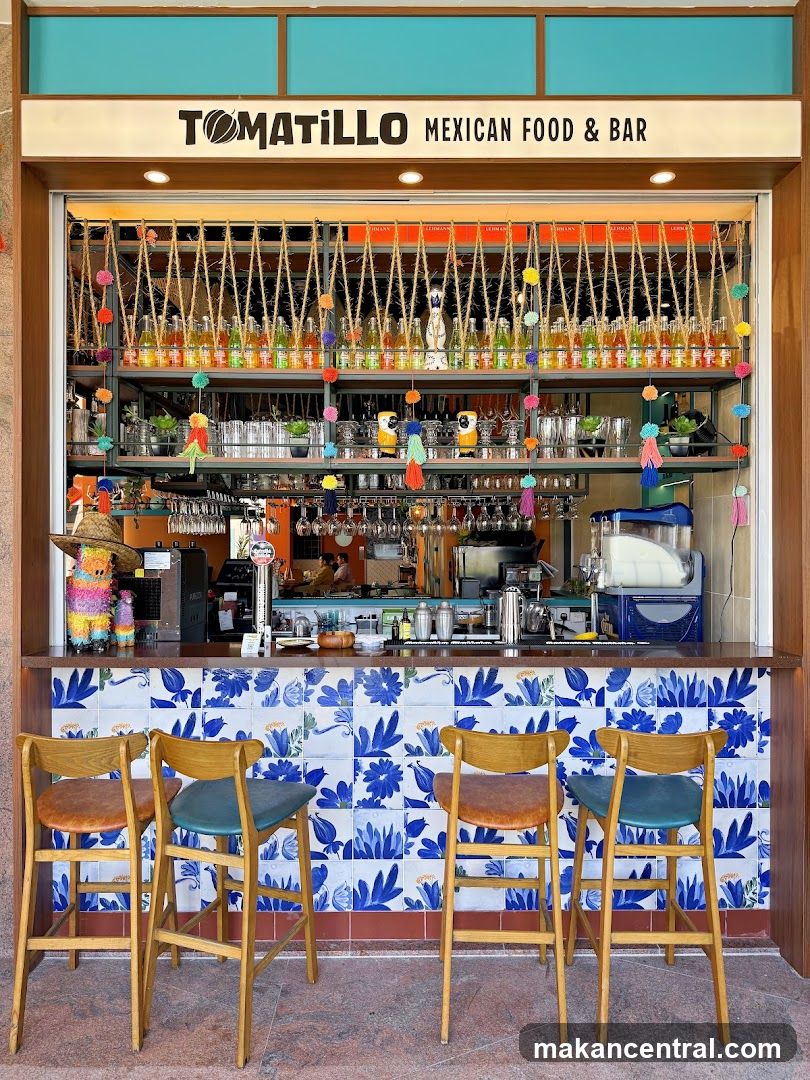 A view of the Tomatillo Mexican Food & Bar storefront, featuring a counter with bar stools, a well-stocked bar area with bottles of alcohol and glassware, and colorful decorations. The restaurant's name and "Mexican Food & Bar" are prominently displayed above the counter.