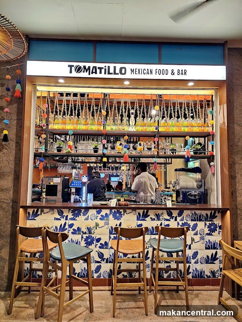 A brightly lit bar area with a sign that reads "TOMATILLO MEXICAN FOOD & BAR". The bar is decorated with blue and white floral tiles and has several bar stools in front of it. Behind the bar, bottles of colorful liquids are displayed on shelves, and two bartenders are visible.