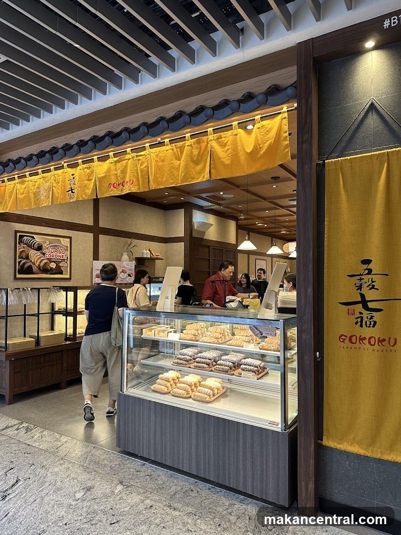 A view of the entrance to Goroku Japanese Bakery, with yellow banners and traditional Japanese architectural elements. Inside, customers are browsing pastries displayed in a refrigerated case.
