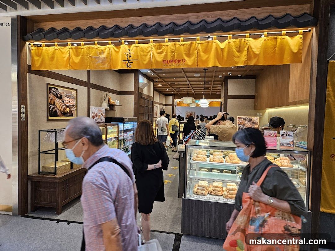 The exterior of a Japanese bakery with a yellow awning and traditional Japanese architectural elements. The shop's name, GOKOKU, is visible on the awning. Inside, display cases are filled with pastries, and customers are browsing.