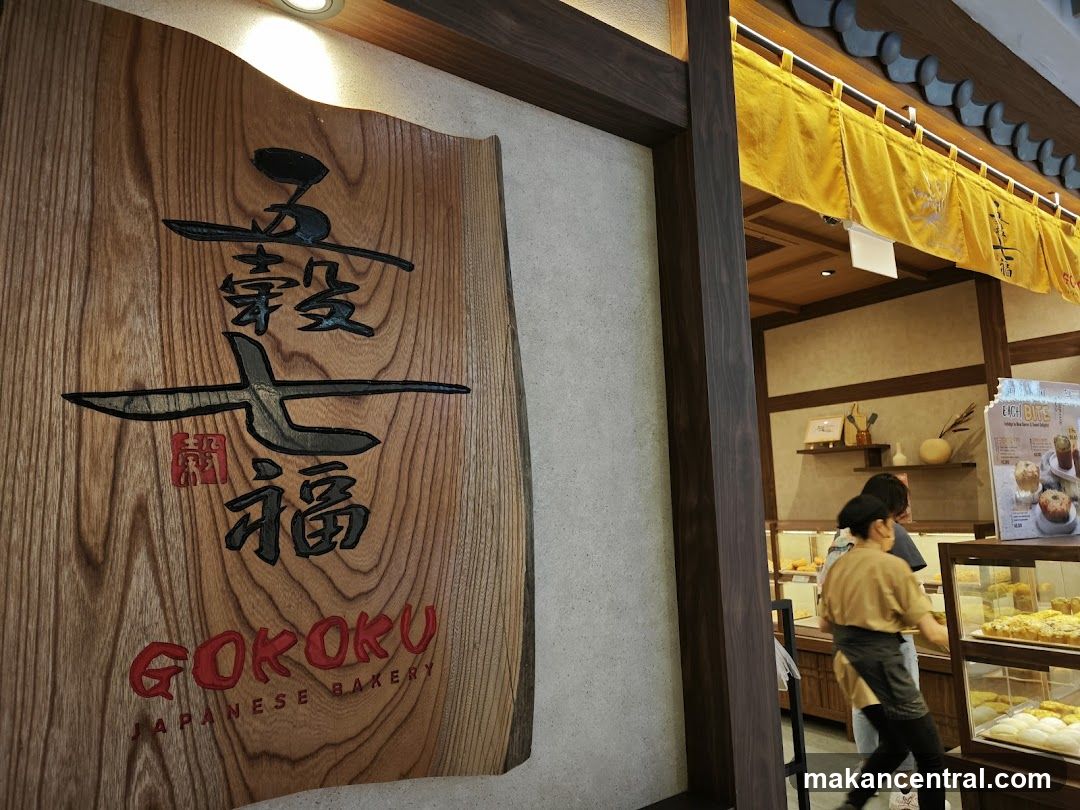 A wooden sign with Japanese characters and the text GOKOKU JAPANESE BAKERY is mounted on a textured wall. To the right, the interior of a bakery is visible, with display cases filled with pastries and a yellow curtain hanging above.