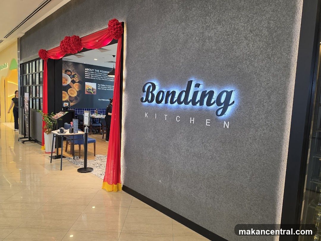 The exterior of a restaurant called Bonding Kitchen. The restaurant's name is illuminated in blue neon lights on a grey textured wall. Red drapes with yellow trim hang around the entrance. Inside, tables and chairs are visible, along with a menu board.