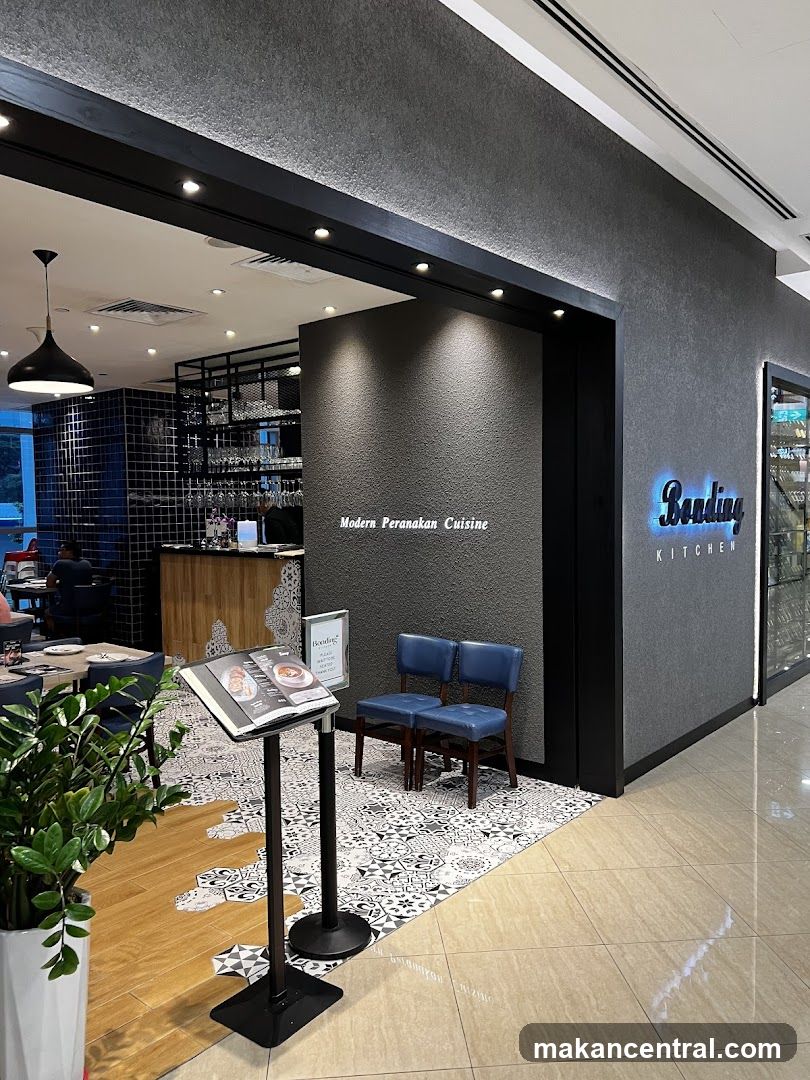 The exterior of a restaurant with a modern Peranakan cuisine theme. The entrance features a dark grey textured wall with the restaurant's name, "Bending KITCHEN," illuminated in blue neon lights. Inside, a bar area with shelves of glassware is visible, along with dining tables and chairs. A menu stand is placed near the entrance.