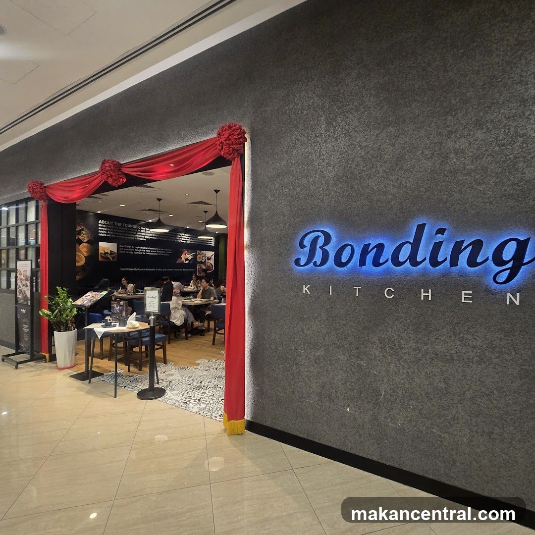 The exterior of a restaurant called Bonding Kitchen. Red drapes frame the entrance, and the restaurant's name is illuminated in blue neon lights on the textured gray wall. Inside, diners are seated at tables.
