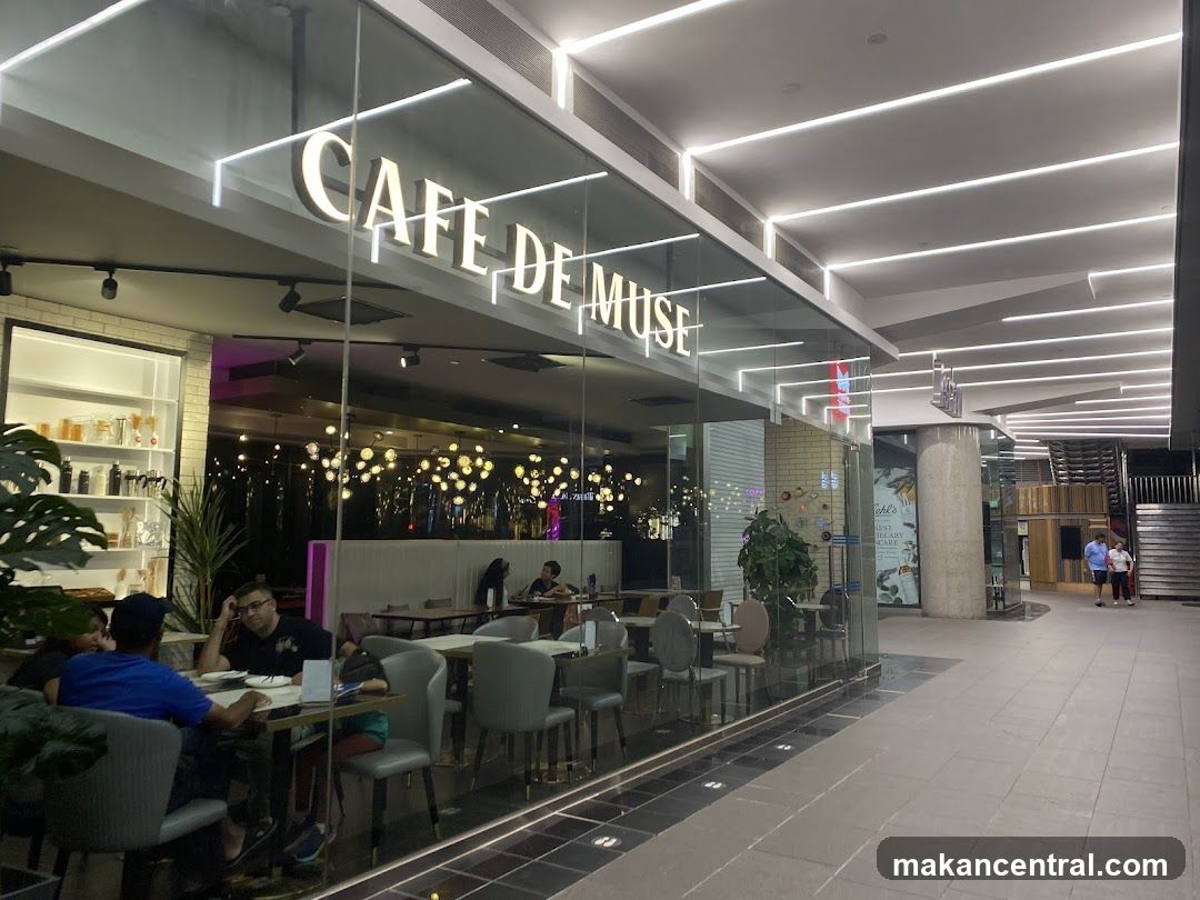 The exterior of a cafe with large glass windows and a prominent sign that reads "CAFE DE MUSE". Inside, tables and chairs are visible, with some patrons seated. The cafe is illuminated by modern ceiling lights and decorative string lights inside.