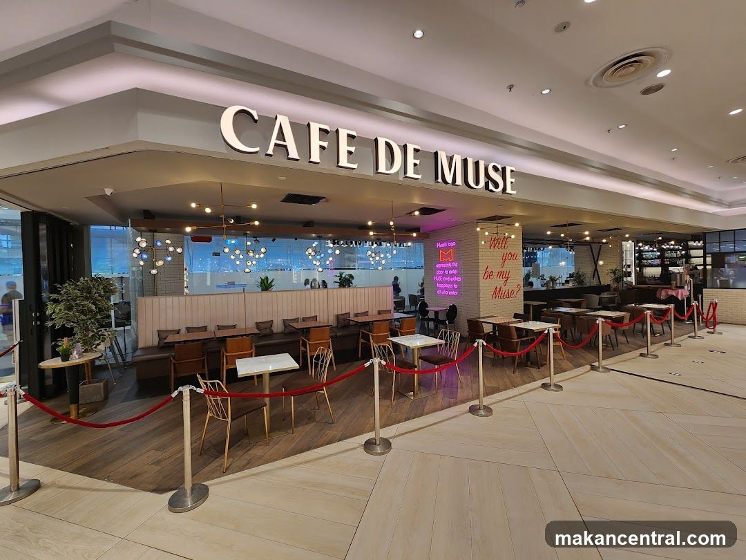 The exterior of a cafe with the name "CAFE DE MUSE" displayed prominently above the entrance. The cafe has an inviting interior with seating areas visible through large glass windows. Red velvet ropes guide customers, and there is a neon sign with the cafe's logo and a tagline.