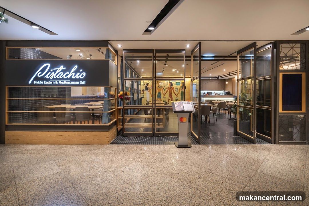 Modern storefront with glass doors and windows, illuminated neon sign reading 'Pistachio' above the entrance. The interior is visible through large glass panels, showing dining tables, decorative wall art of figures in traditional attire, and a counter area. A standing menu kiosk with Mastercard logo stands at the entrance. The floor is polished stone tile.